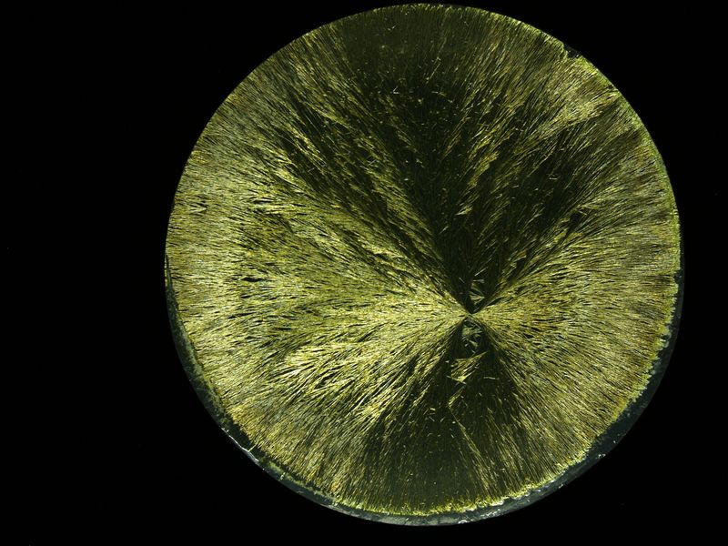 Mikroskopische Ansicht eines polierten und geätzten metallischen Materials mit nadelartigen Kristallstrukturen, die radial von der Mitte des runden Schnitts ausgehen.